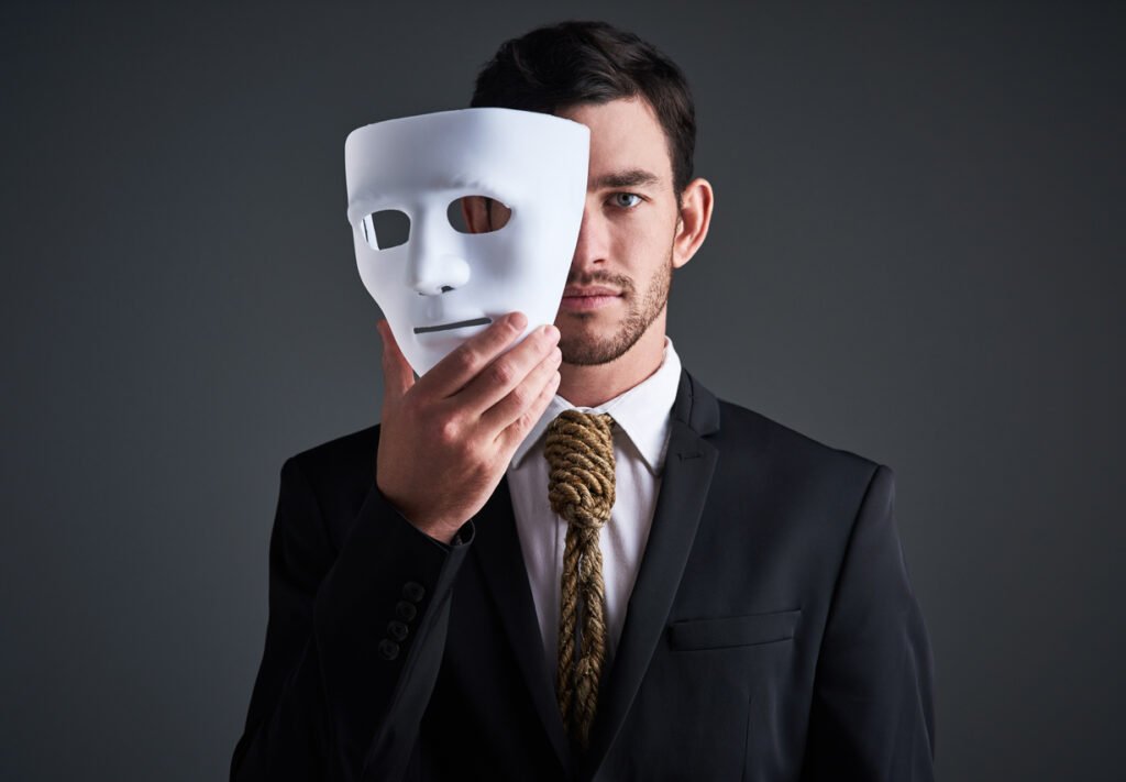 Man in a suit holding a white mask over half of his face, revealing one side of his real face while the other is concealed, symbolizing hidden identity or dual persona.